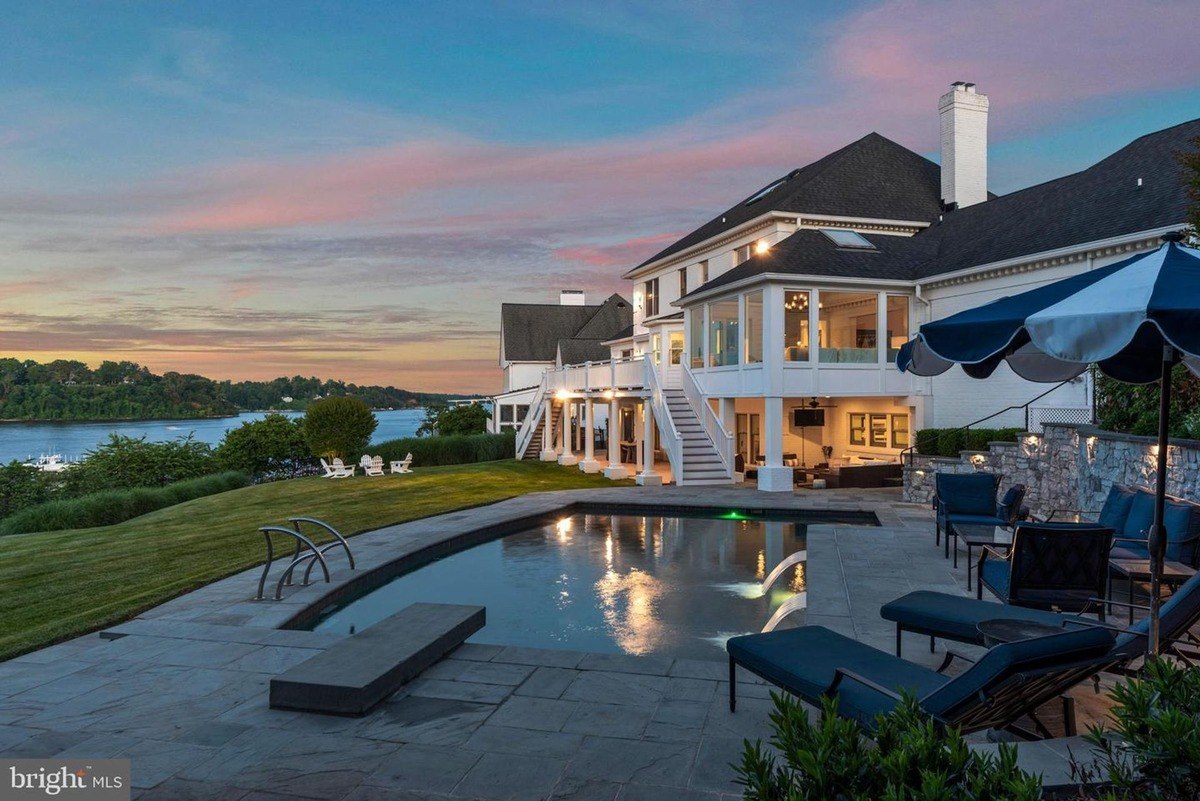 Large waterfront home has a pool and patio furniture at sunset.