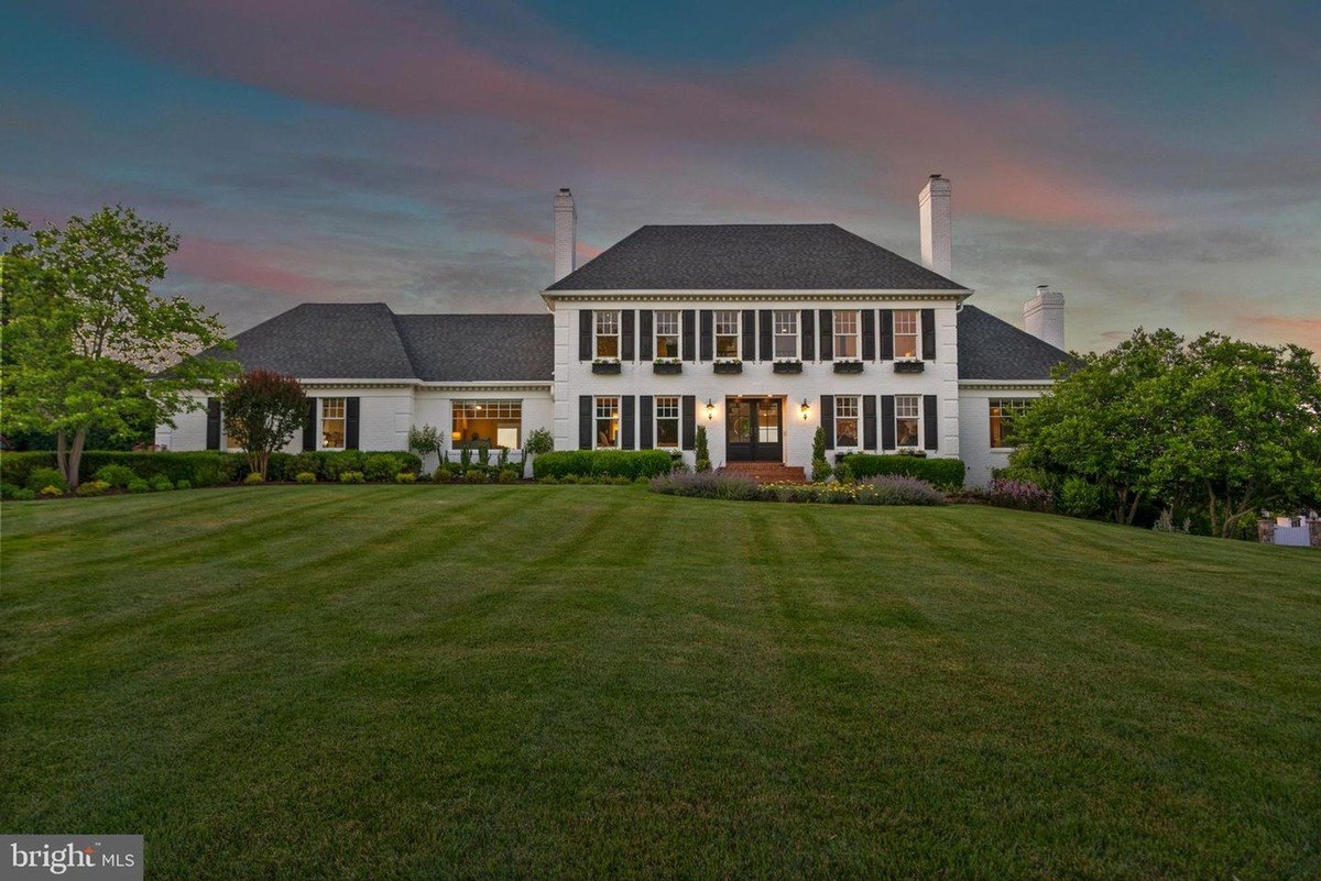 Large white house has black shutters and is set on a large lawn at dusk.