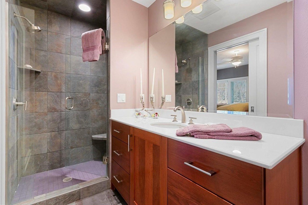 Wood cabinets and a white countertop complement the pink walls and slate-tiled glass shower in this bathroom.