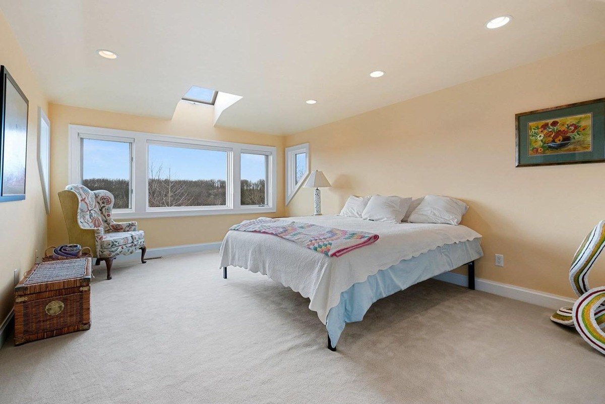 Bedroom has beige walls, carpeted floor, large windows with a view, and a bed with a quilt.