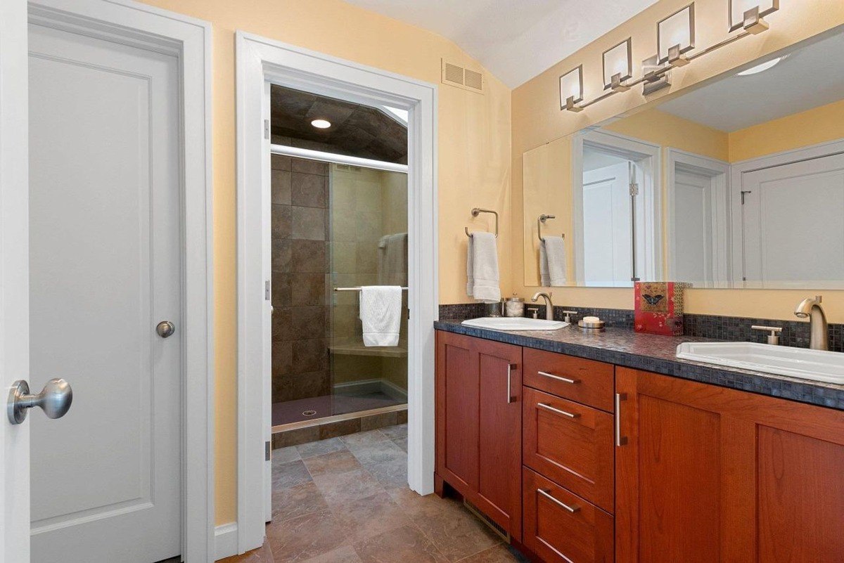 Bathroom has double sinks, wood cabinets, and a shower with stone tile.