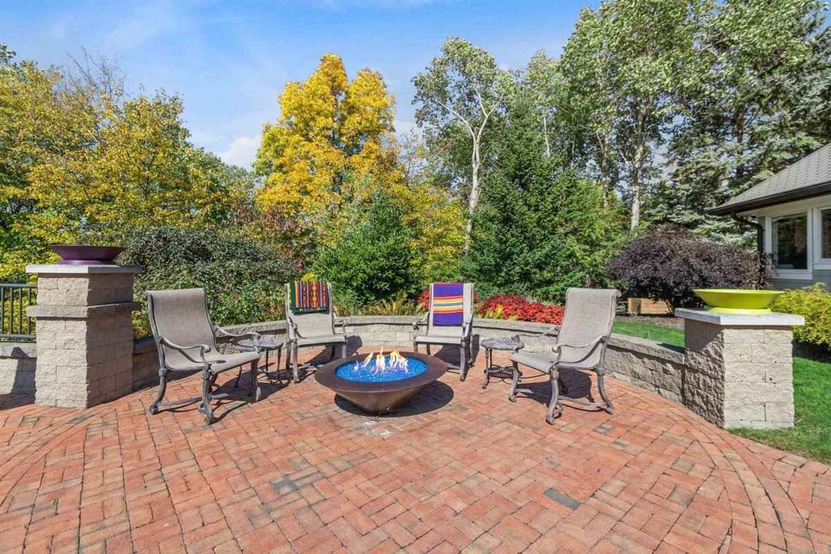 Patio has brick flooring, fire pit, and chairs surrounding it, with trees in the background.