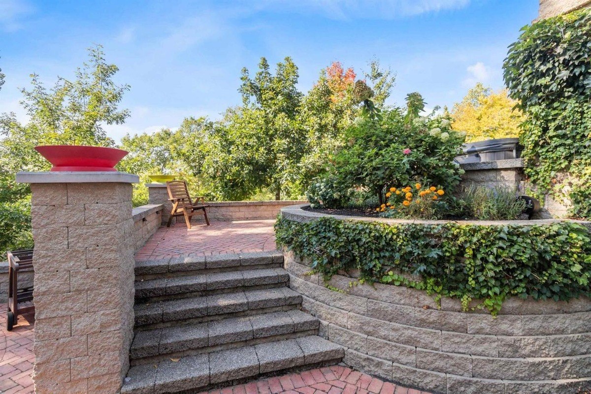 The brick patio includes a retaining wall with greenery, steps, and a chair.