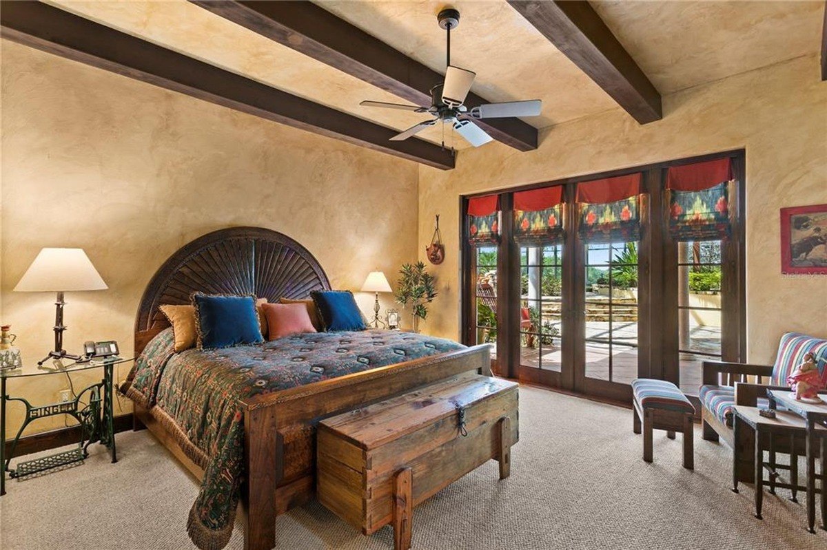 Bedroom features a large wooden bed with a unique headboard, exposed wooden beams, and French doors leading to a patio.