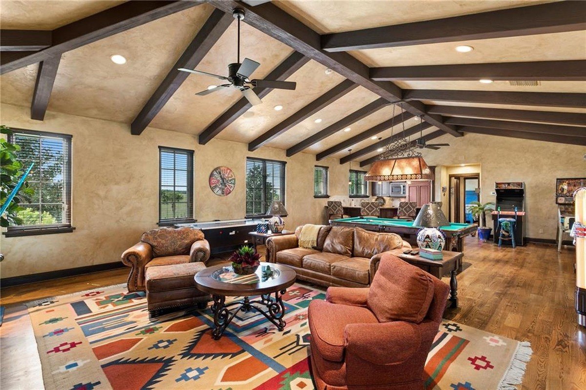 Game room features a pool table, seating area with a large rug, and exposed wooden beams in a high-ceilinged space.