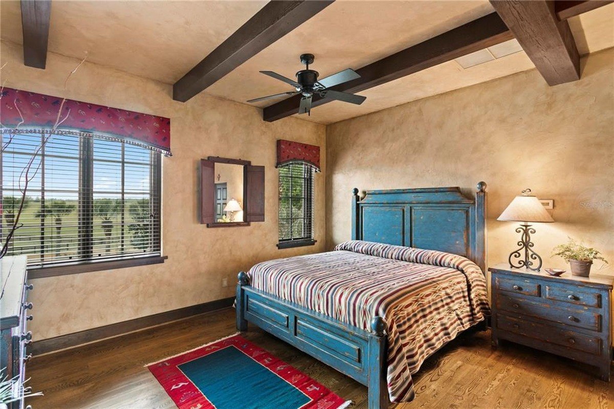 Bedroom features a blue wooden bed with a striped comforter, exposed wooden beams, and a view of a landscape.