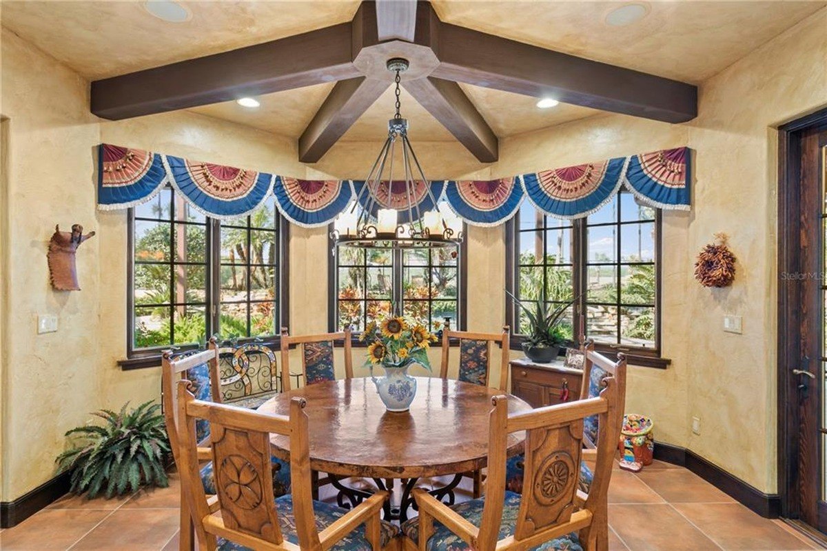 Dining area features a large round table surrounded by intricately carved chairs, a chandelier, and windows with decorative valances.