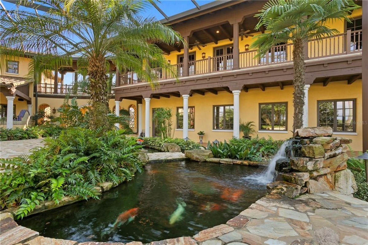 Freeform pool with a rock waterfall feature is nestled in a lush tropical garden, surrounded by a Mediterranean-style house with a covered patio.