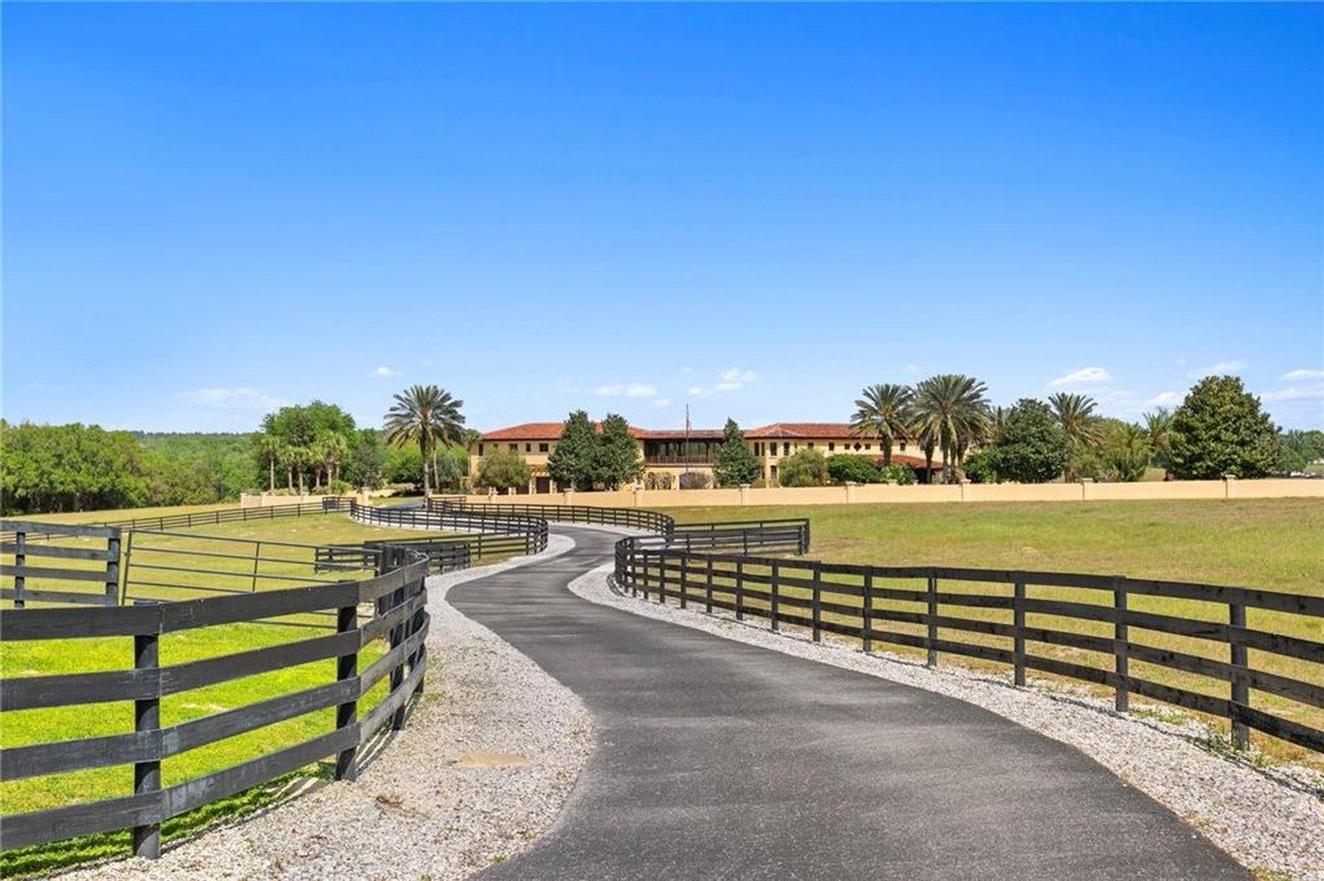 Long driveway winds through a grassy field, leading to a large Mediterranean-style house.