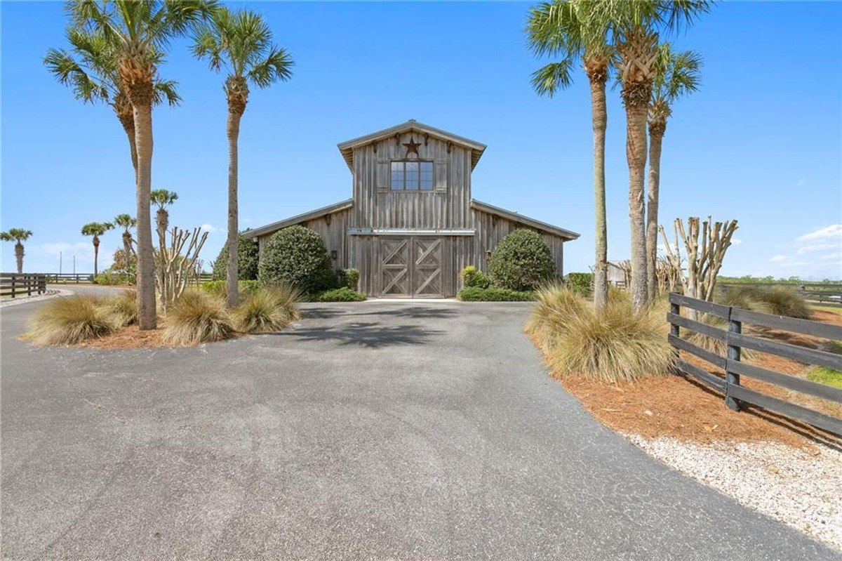 Large wooden barn with double doors sits in a sunny field, surrounded by palm trees and a fence.