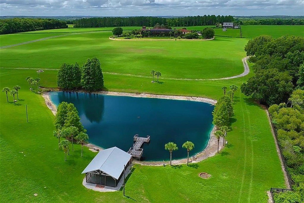 Aerial view shows a large pond with a dock and a screened-in pavilion, situated on a sprawling green property with a house in the distance.