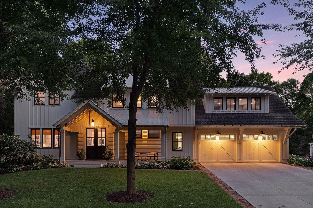 Exterior at dusk a modern farmhouse with illuminated windows, a covered porch, and a two-car garage.