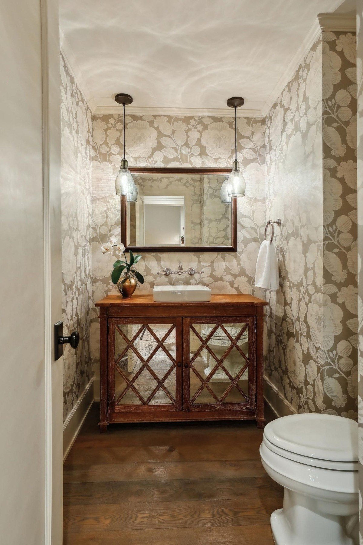 A cozy powder room with floral wallpaper, a wooden vanity with mirrored panels, and pendant lights.