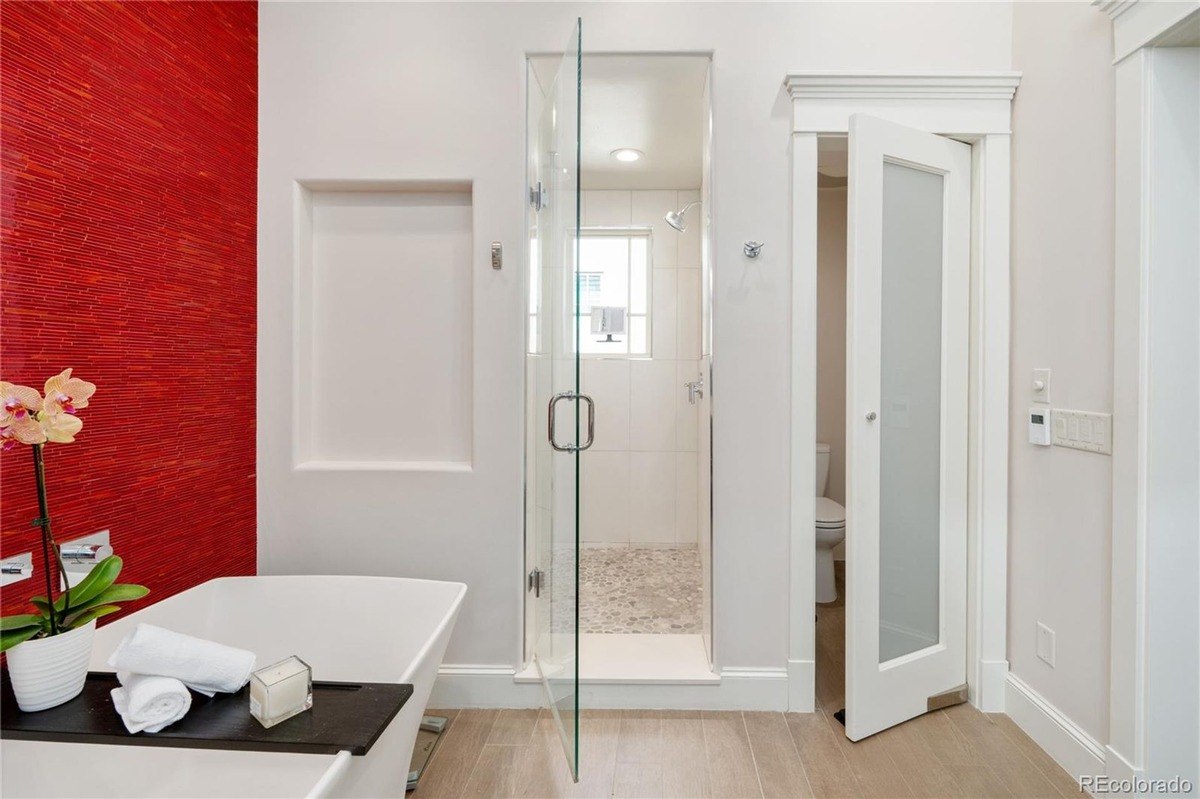 Bathroom has red wall, freestanding tub, glass shower enclosure, and toilet in separate room.