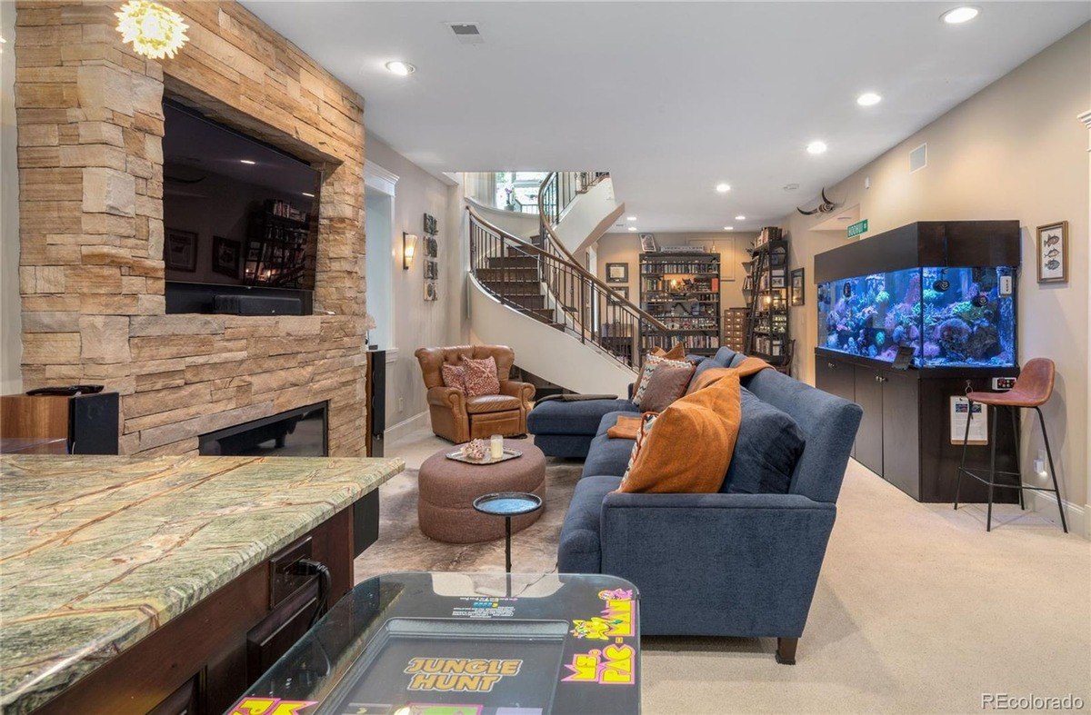 Game room has stone fireplace with TV, sectional sofa, large aquarium, and spiral staircase.