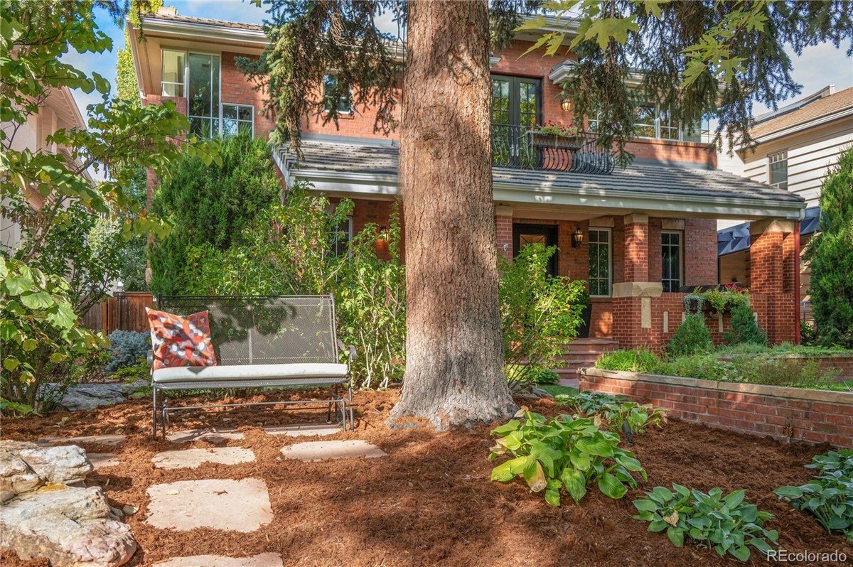 Brick house has large tree in front, landscaping with mulch, and metal bench.