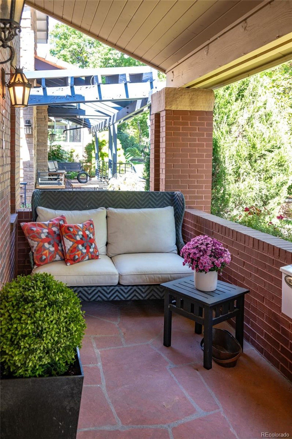 Porch has wicker settee with pillows, small table, and potted plant.