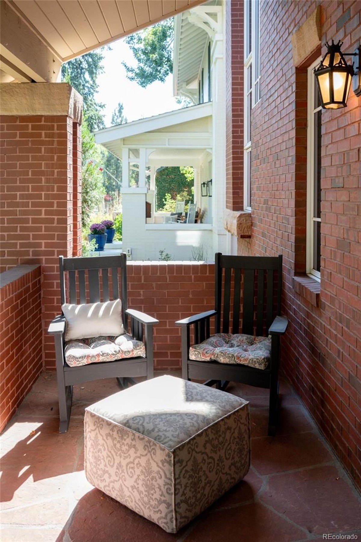 Brick porch has two rocking chairs, ottoman, and light fixture.