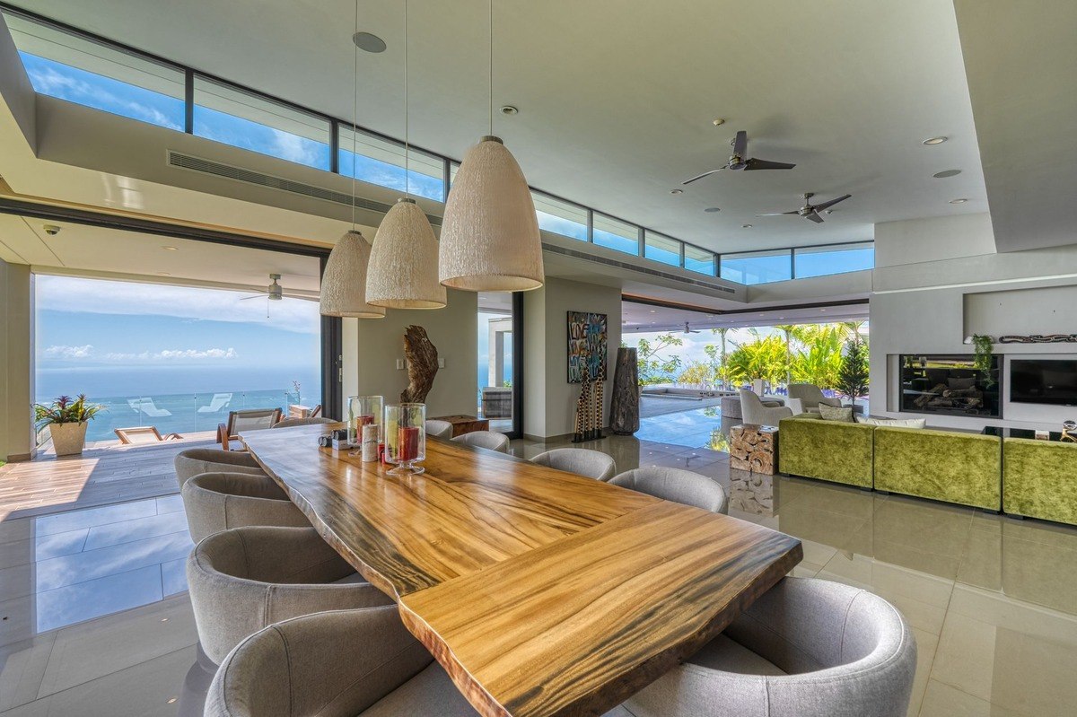 Dining area has long wood table, ocean view, and modern lighting.