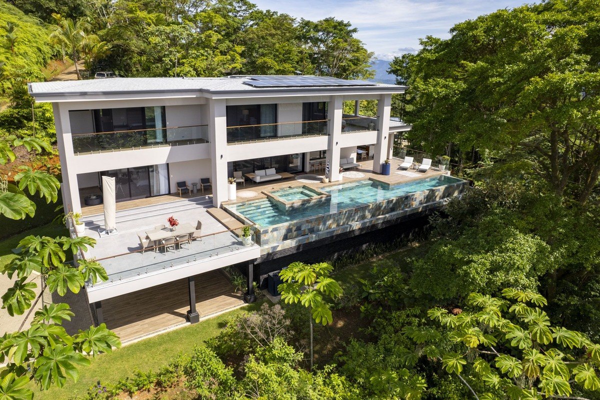 Aerial view of a luxurious, modern two-story home with a large infinity pool and expansive deck nestled amidst lush green tropical foliage.