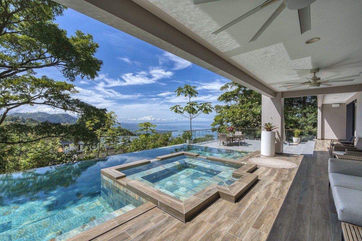 From a covered patio, a view of an infinity pool and hot tub extends to a breathtaking vista of ocean and mountains under a bright blue sky.