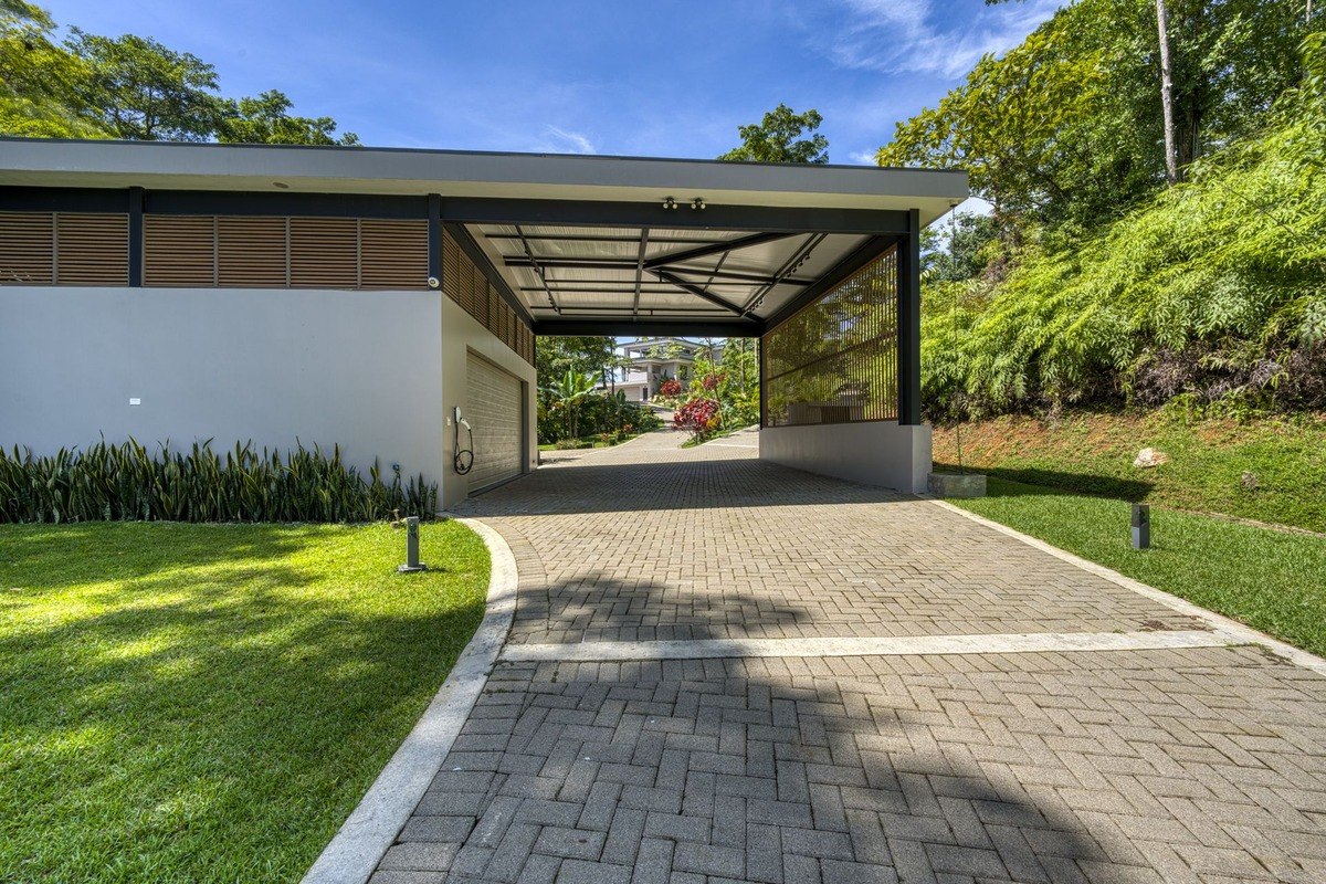 Modern carport leads to a curving brick driveway, set against a backdrop of lush tropical landscaping.