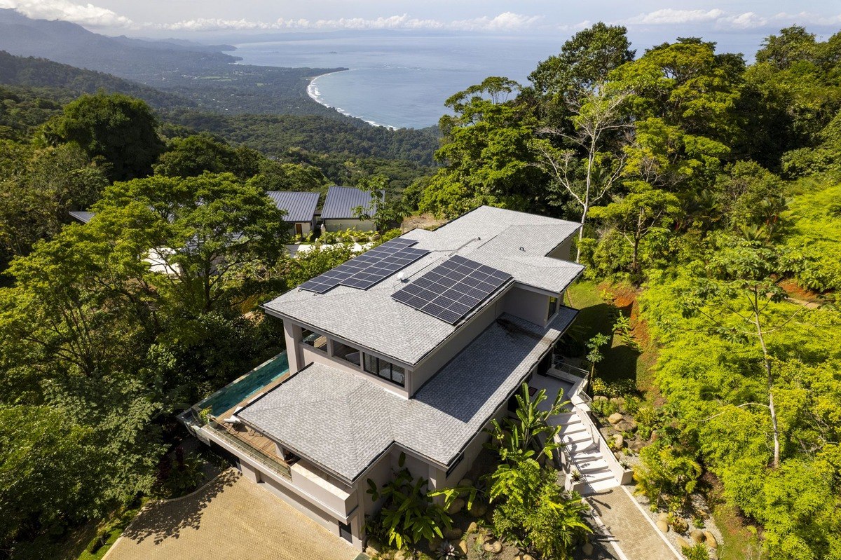 An aerial view shows a modern, solar-powered house with a pool overlooking a stunning ocean and mountain vista.