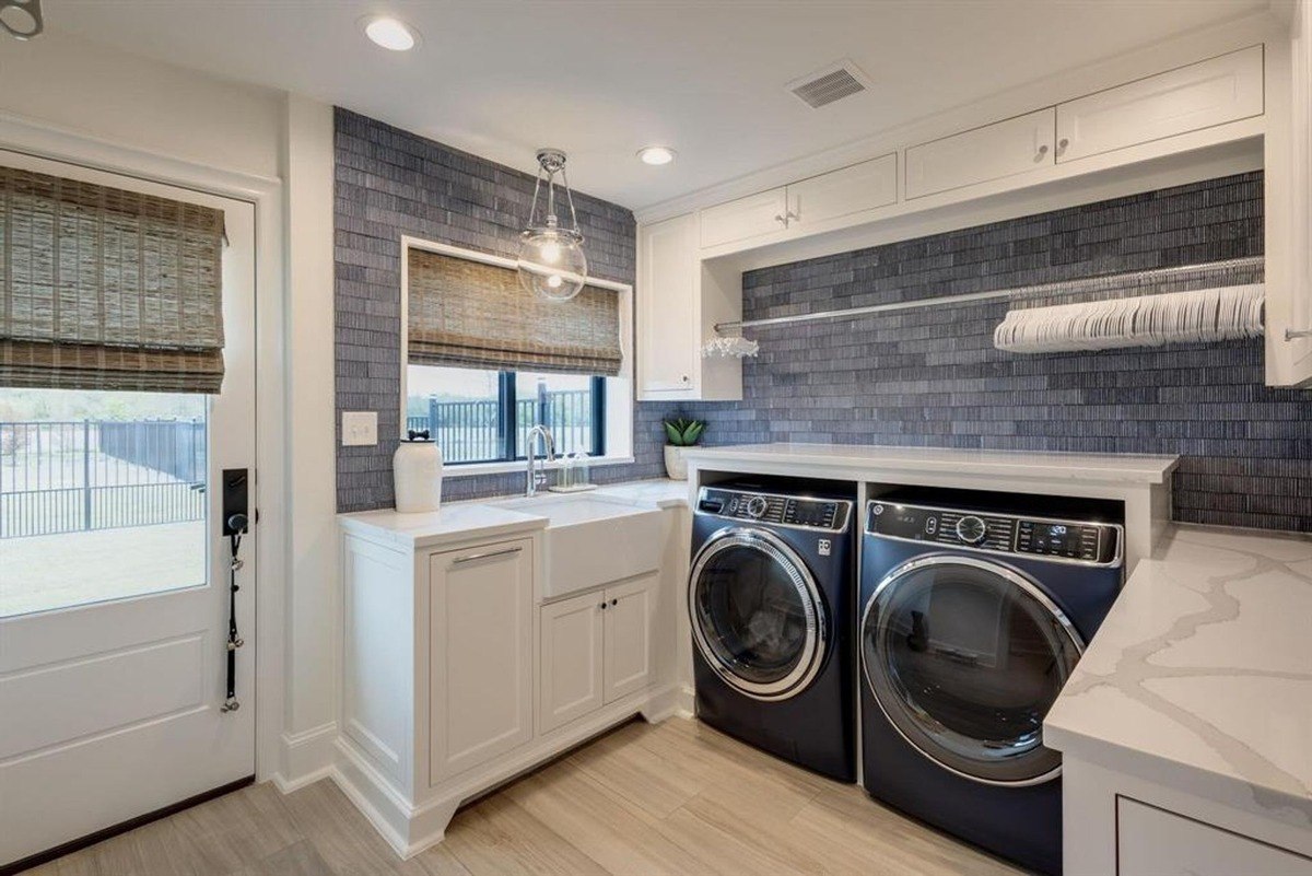 Laundry room includes a deep farmhouse sink, a set of dark blue front-load machines, and a hanging rod under cabinets.