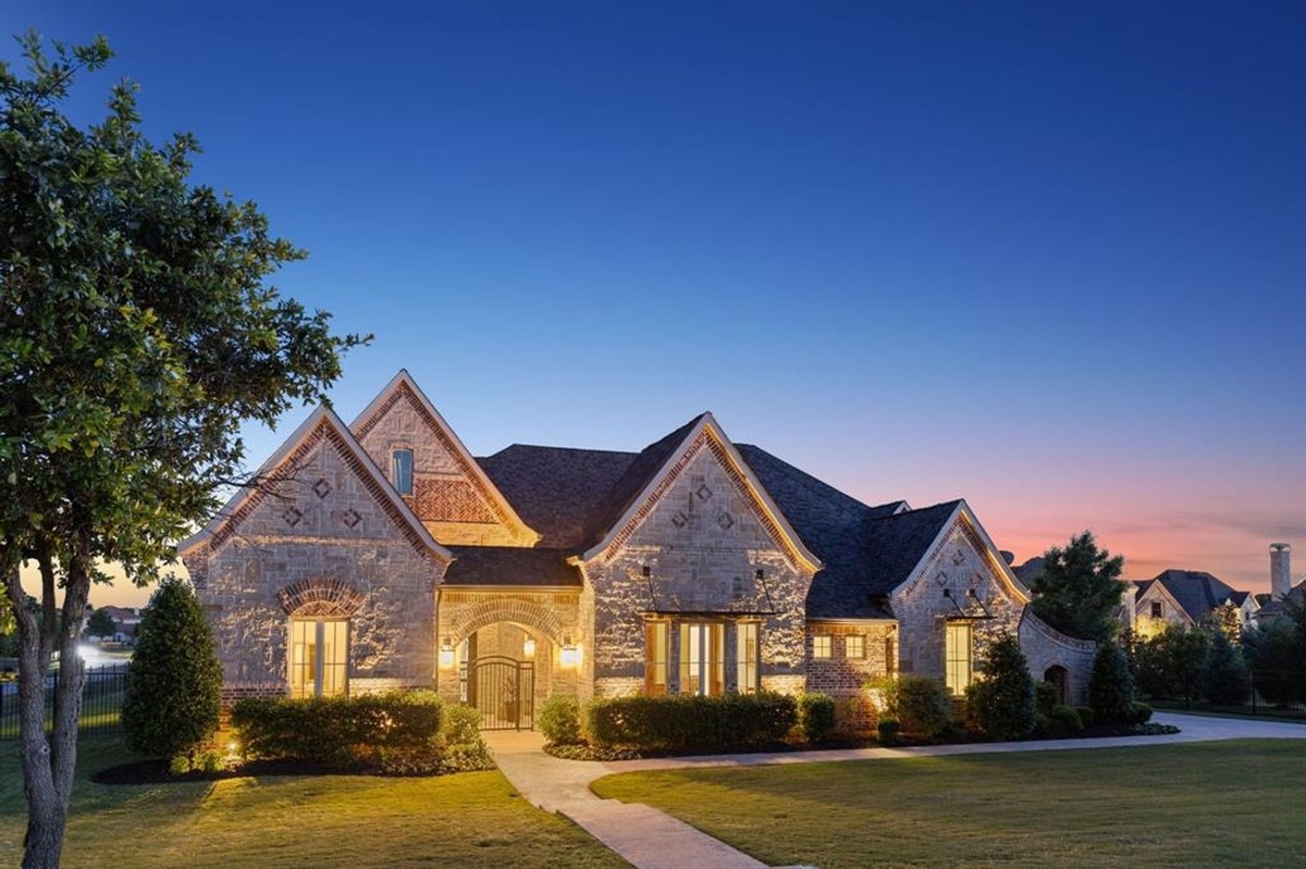 Exterior of a stone house with a front yard at sunset, featuring multiple peaked roofs and warm lighting.