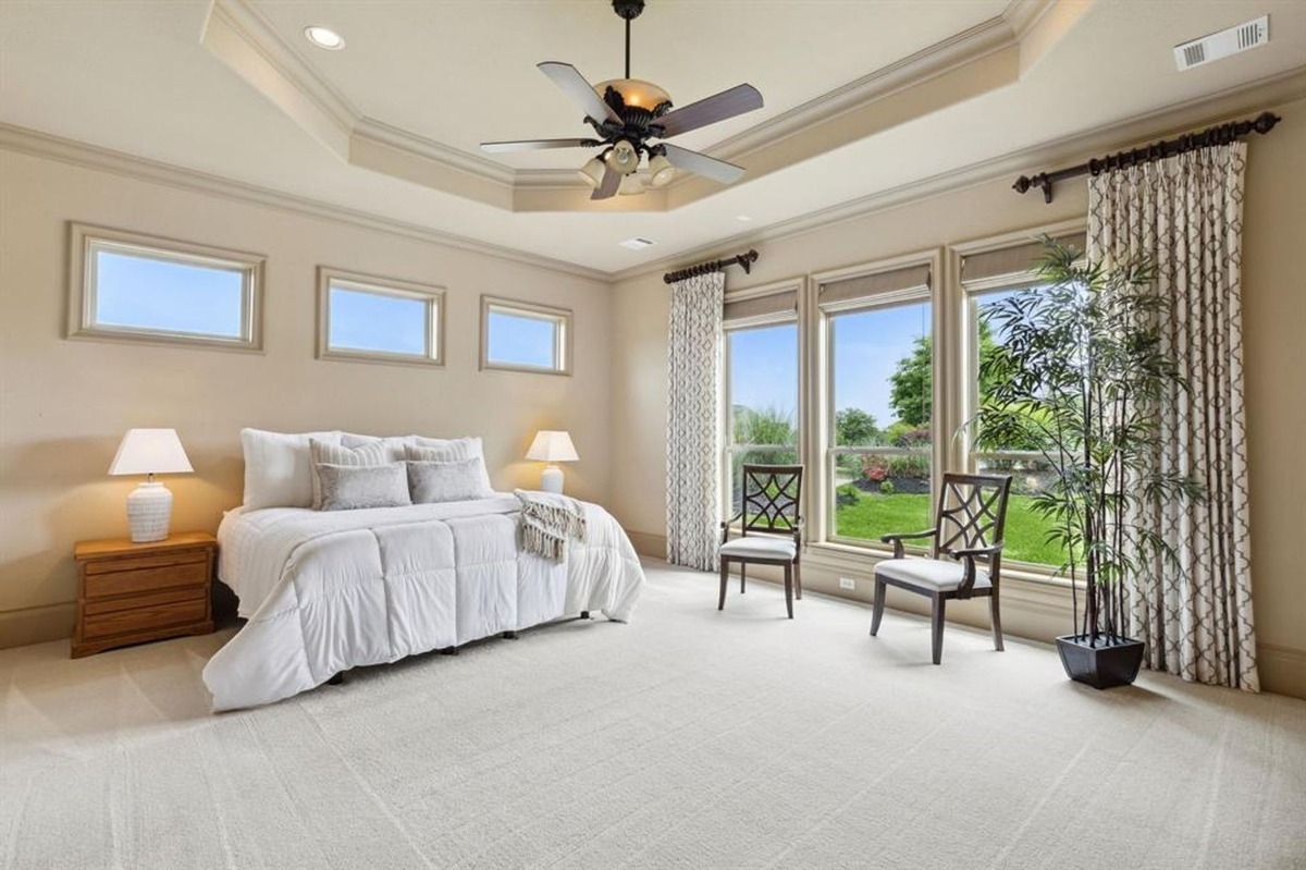 Bedroom with a tray ceiling, large windows, and neutral decor overlooking a landscaped backyard.