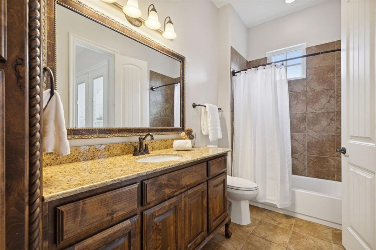 Bathroom with a granite-topped vanity, tiled shower-tub combination, and ornate mirror frame.