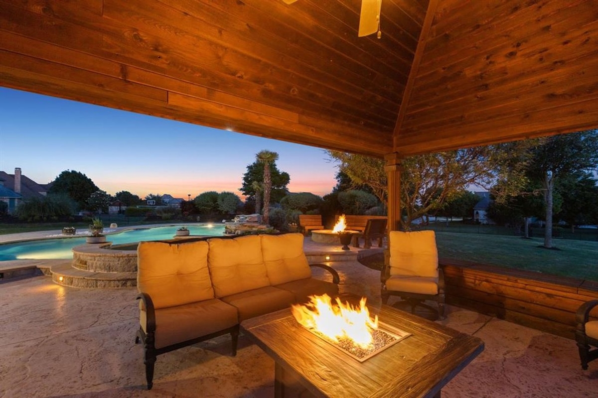 Cozy seating area under a wooden pergola with a fire pit in the foreground overlooking a serene backyard pool.