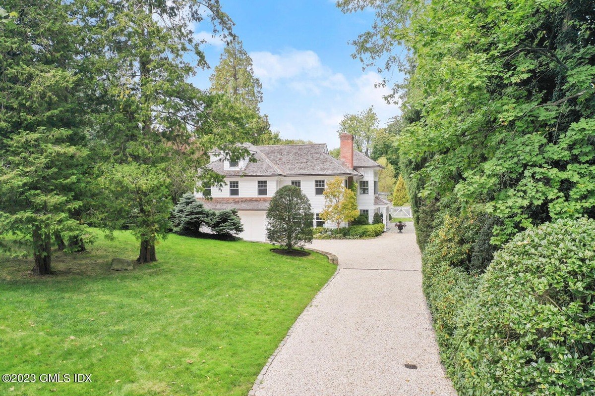 Large white house has a long driveway and is surrounded by green trees.