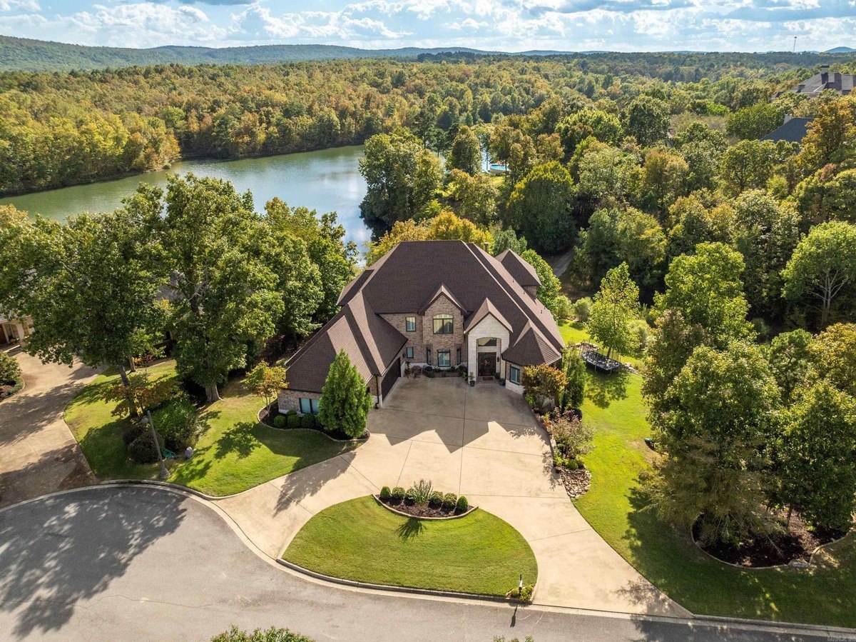 Large house with brown roof sits on a large lot near a lake, surrounded by trees and hills.