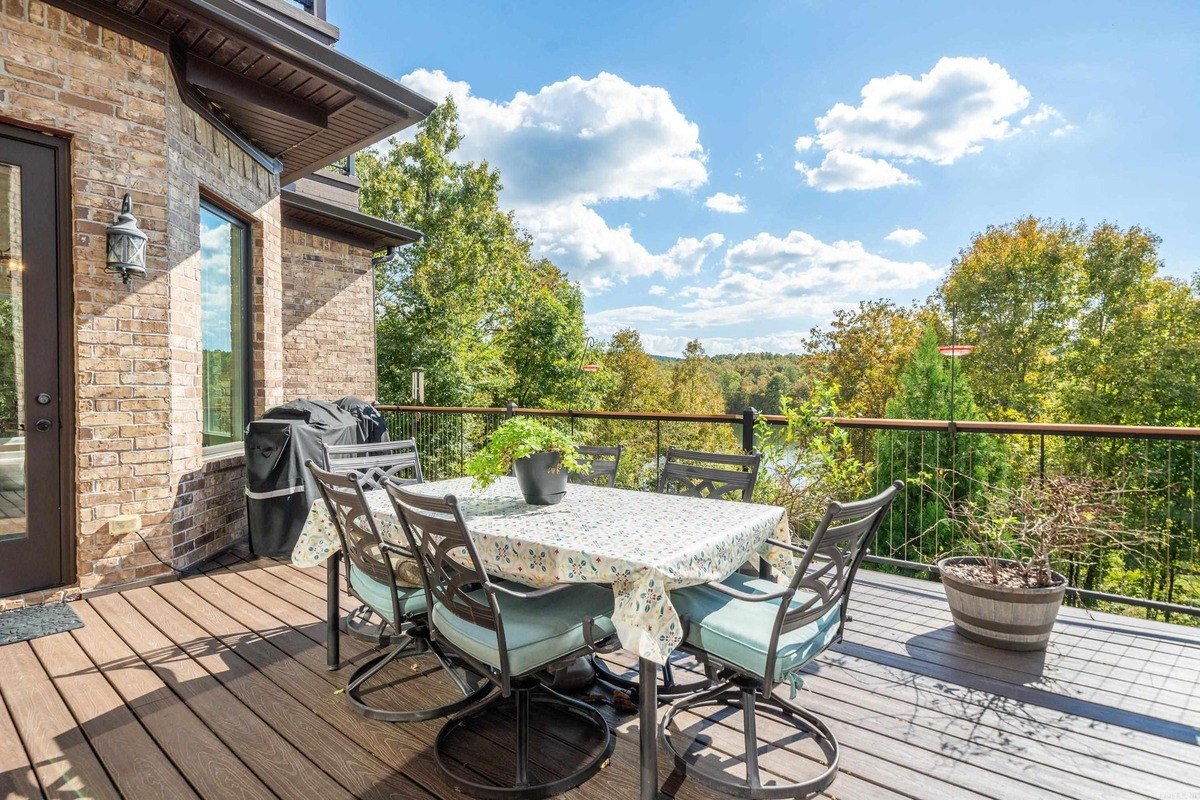 Outdoor dining area with a table and chairs on a deck overlooking a wooded area and lake.