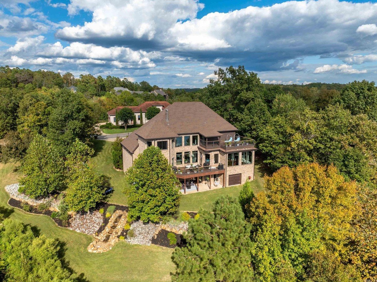 Large brick house with a deck overlooks a wooded area under a partly cloudy sky.