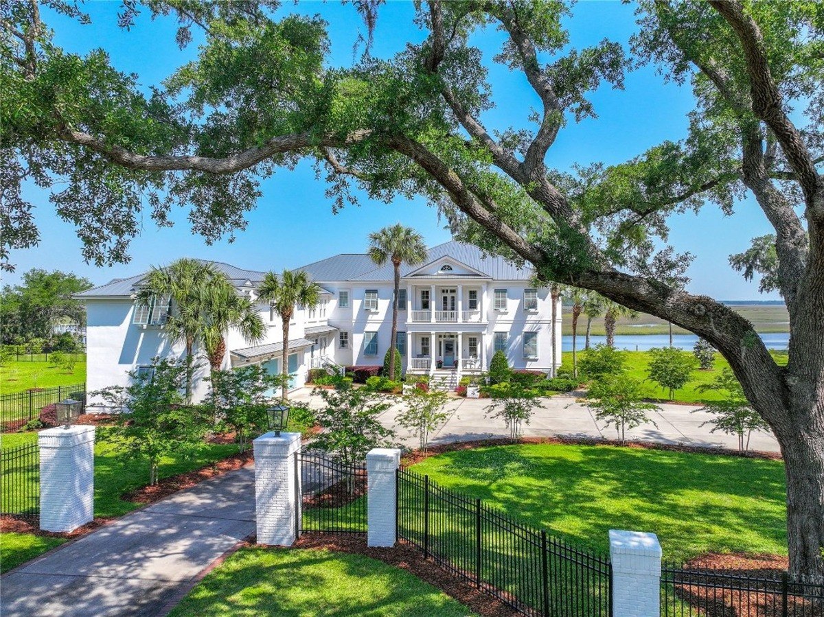 Large white house with a wrap-around porch is shown, situated on a waterfront property with lush landscaping.