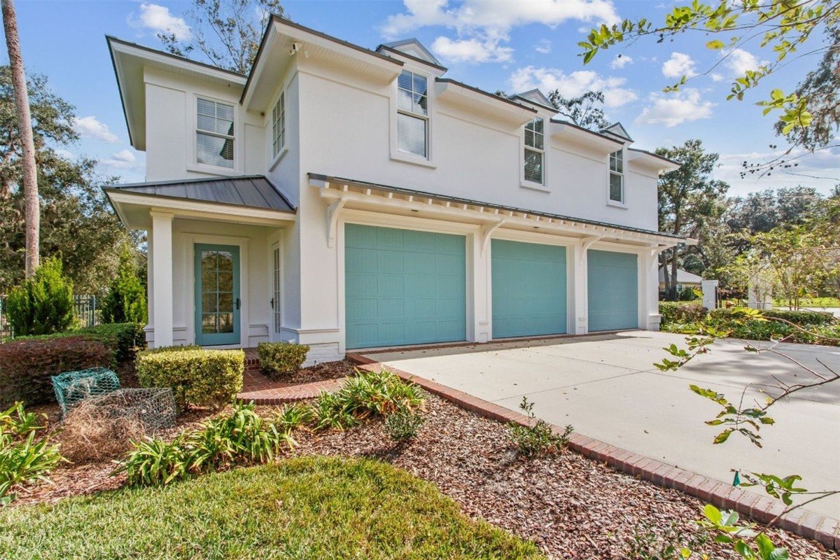 White house with three light teal garage doors is shown, surrounded by landscaping.