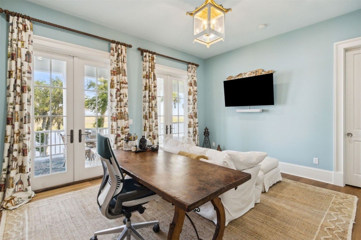 Office has a large wooden desk, white slipcovered couch, and French doors with a water view.