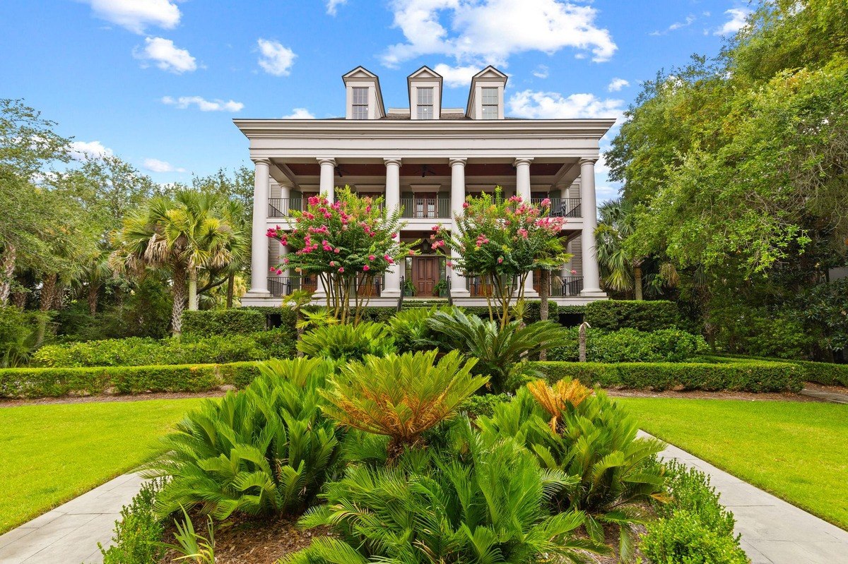 Large white mansion with columns and balconies is surrounded by lush landscaping, including palm trees and flowering shrubs.