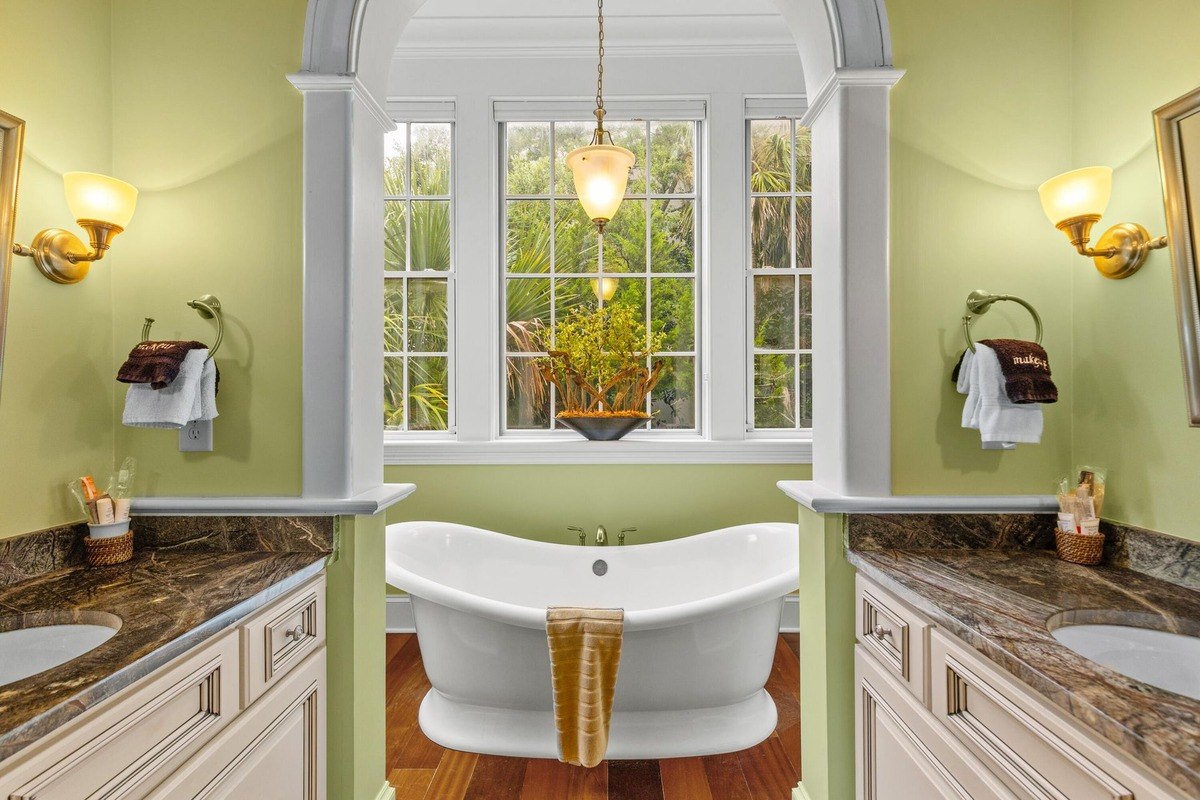 Freestanding bathtub sits between double vanities in a bathroom with light green walls and large windows.