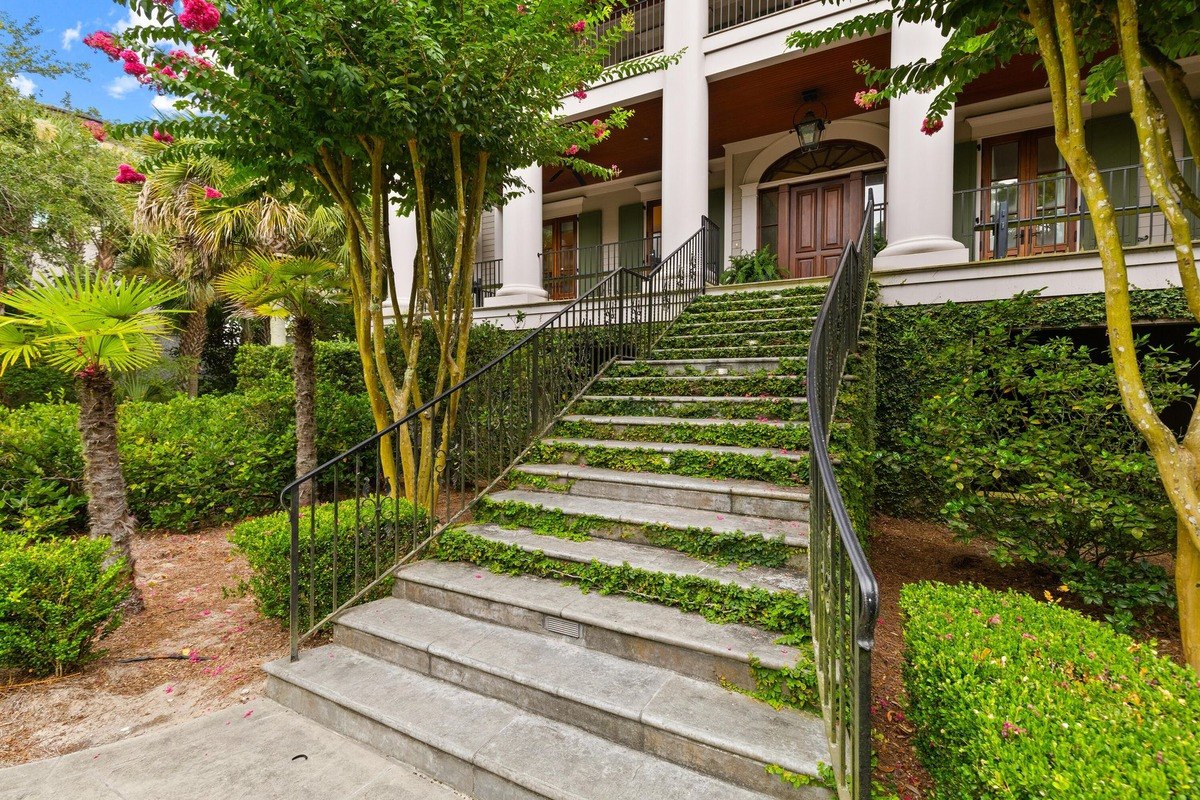 Stone steps leading to a large house's entrance are lined with greenery and flanked by lush landscaping.