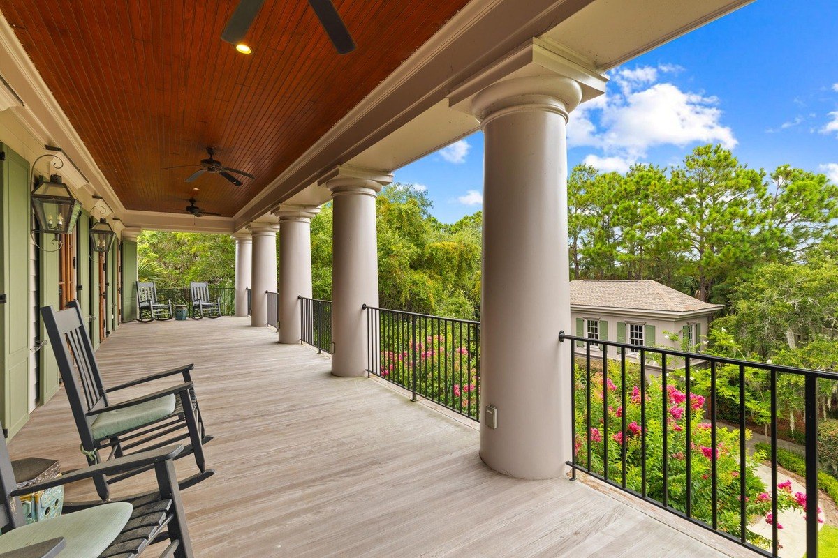 Porch has columns, a wooden floor, rocking chairs, and overlooks a garden and houses.