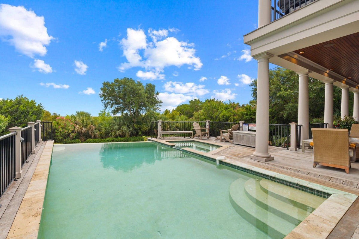 Swimming pool is surrounded by a deck, columns, and has a view of lush greenery.