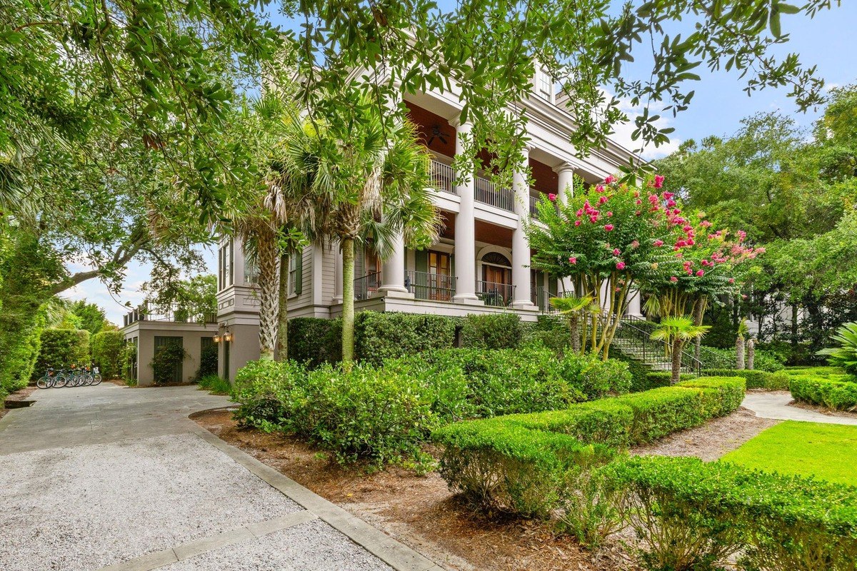 Large, light-colored house with columns and a wrap-around porch is surrounded by lush landscaping.