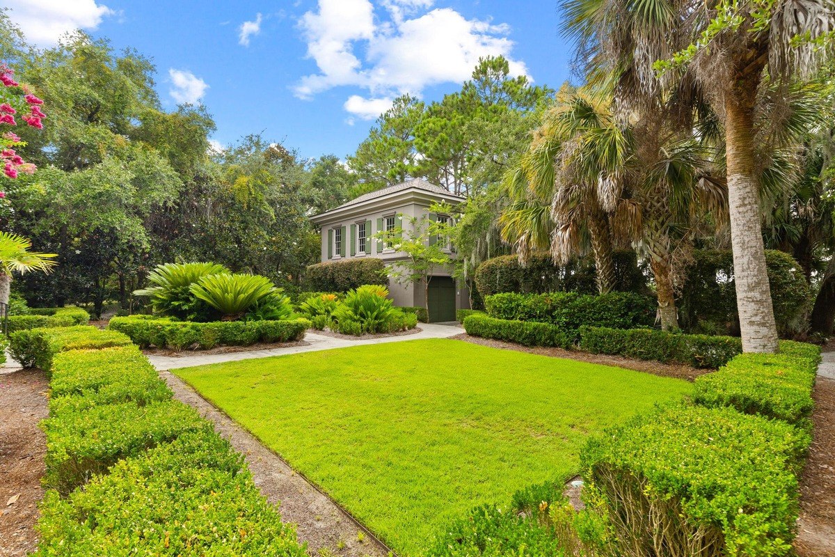 House with green shutters is surrounded by manicured lawns and tropical plants.