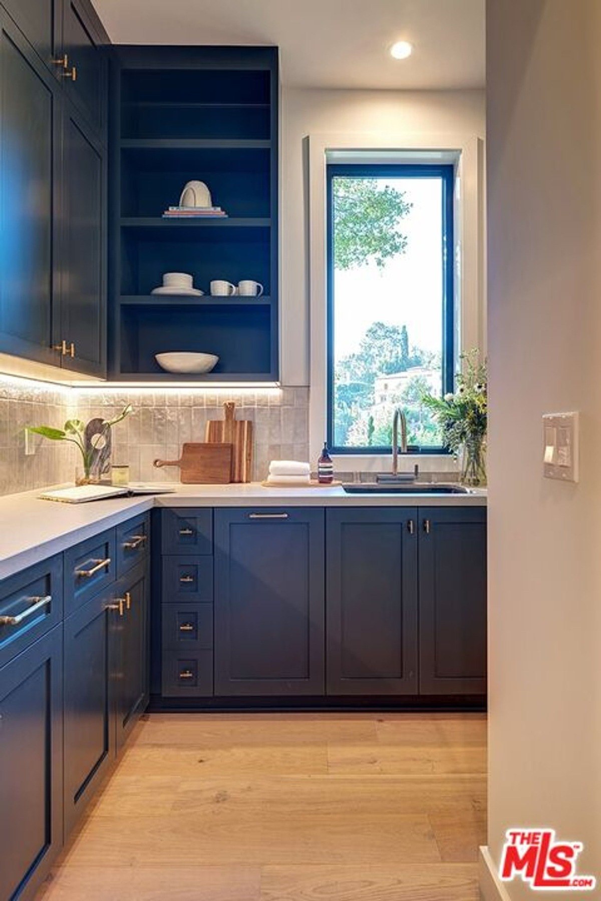 Kitchen pantry features deep navy cabinets with brass handles, open shelving, and under-cabinet lighting alongside a bright window view.