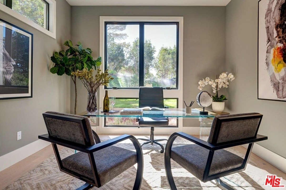 Home office offers a glass desk, cozy seating, wall art, and a large window that frames lush greenery.