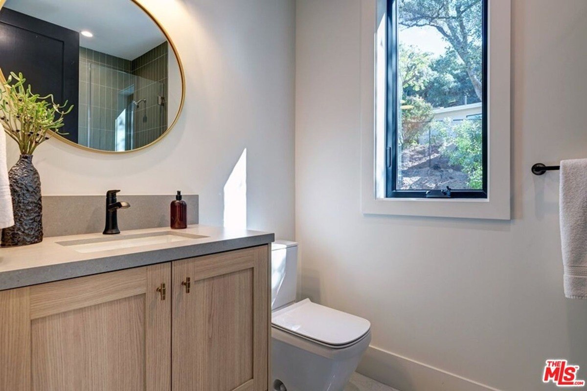 Circular mirror above a light wood vanity with a modern black faucet near a window that overlooks the greenery outside.