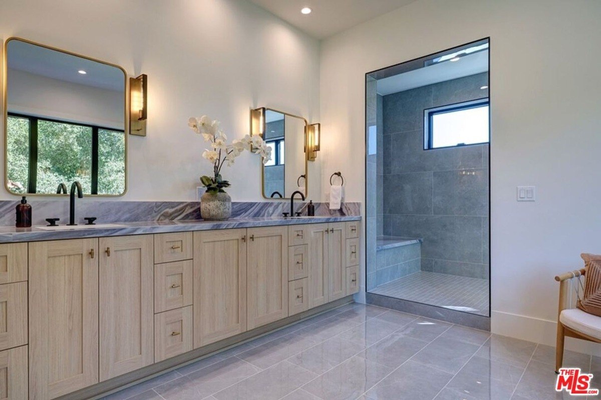 Double vanity area includes light wood cabinets, dual mirrors with brass sconces, and a glass-enclosed walk-in shower.